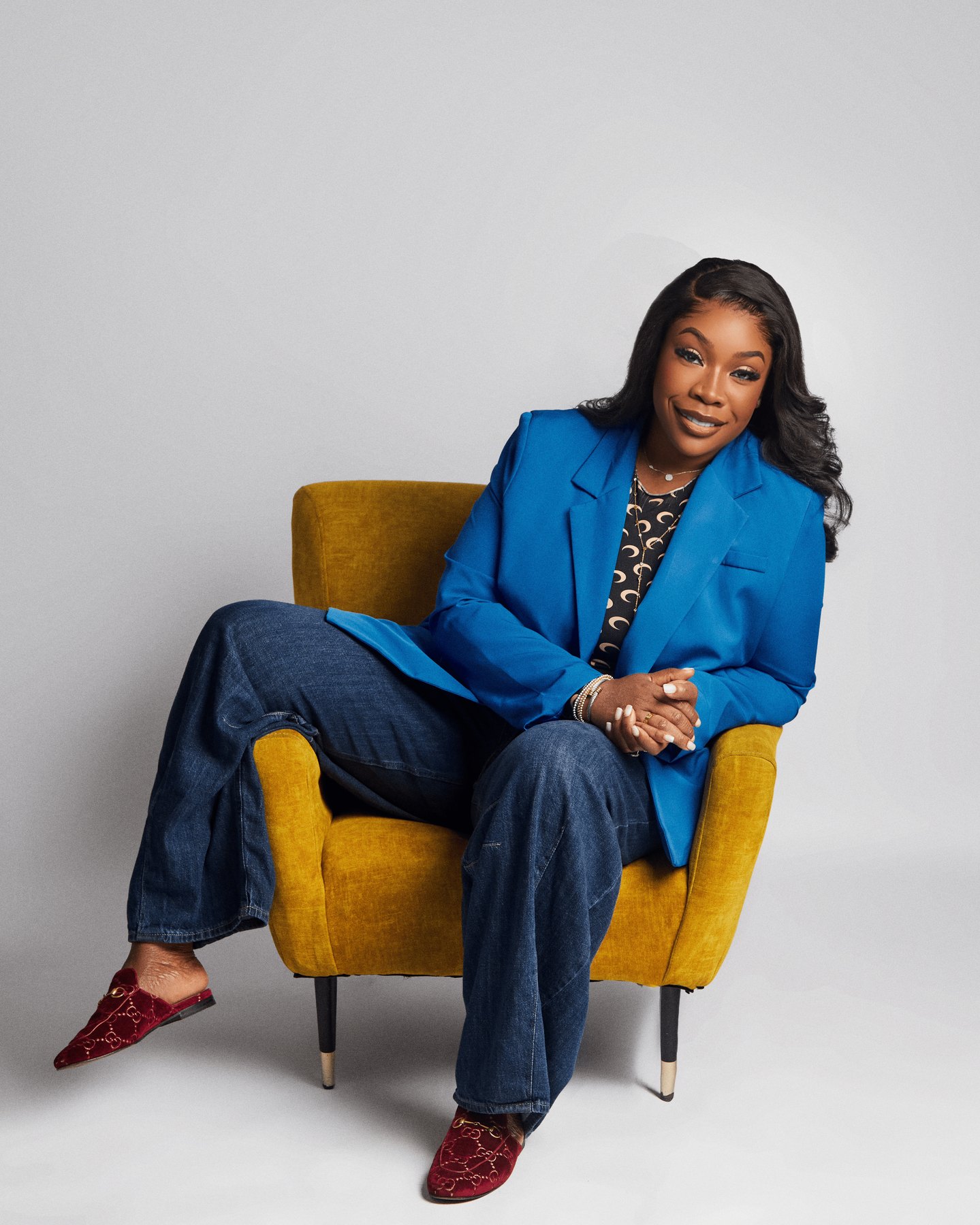 Dr. Bukky George-Taylor seated in a mustard velvet chair, wearing a blue blazer and jeans.